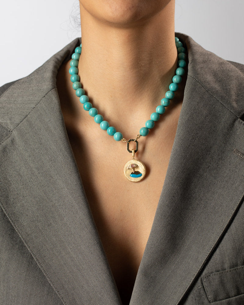 Person wearing a turquoise beaded necklace with a gold pendant against a neutral background