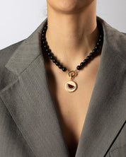 Person wearing a black beaded necklace with a gold pendant, set against a neutral background.